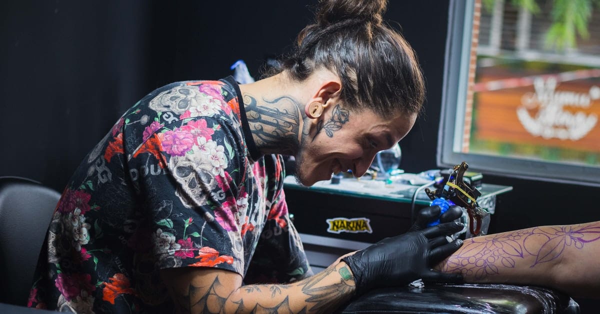 tattoo artist with lots of tattoos smiling as he fills out a tattoo on a womens arm