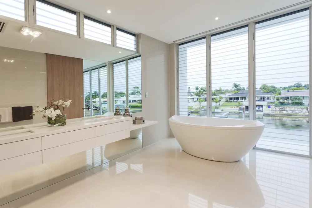 a spacious white bathroom design with floor to ceiling jalousy window, light wood accent wall and white acrylic freestanding tub